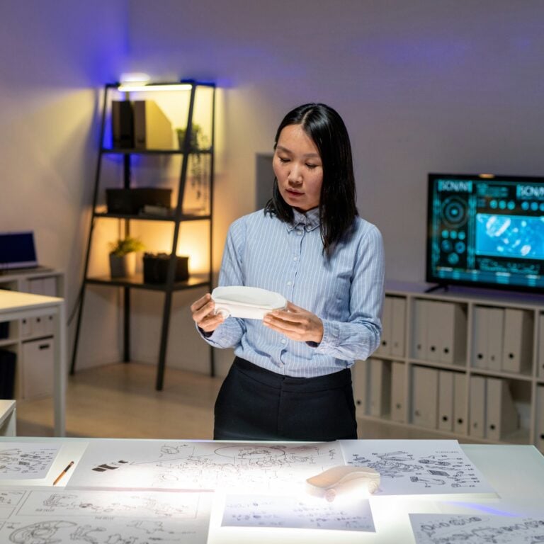 A woman stands at a desk covered with technical drawings and papers, holding a 3D-printed object. Shelves with plants and a digital display screen are visible in the background, suggesting a modern office or lab setting.