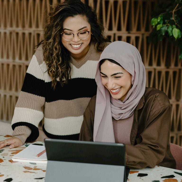 Two women smile while looking at a laptop. One is wearing glasses and a striped jumper, and the other wears a hijab and brown shirt. They appear to be working or studying together at a table indoors.