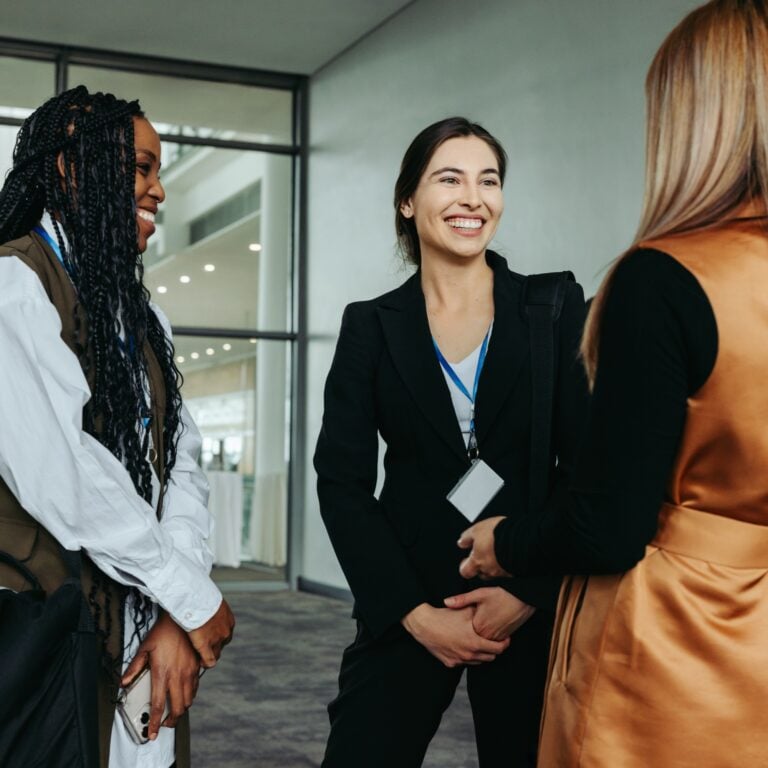Three women, dressed in professional attire with conference badges, stand indoors in conversation. One woman in a black suit smiles warmly whilst the others listen, suggesting a friendly networking event.