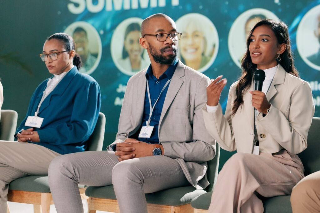 Three professionals sit on a panel at a summit. One woman holds a microphone and speaks whilst the others listen. A summit banner with blurred images and text is visible in the background.
