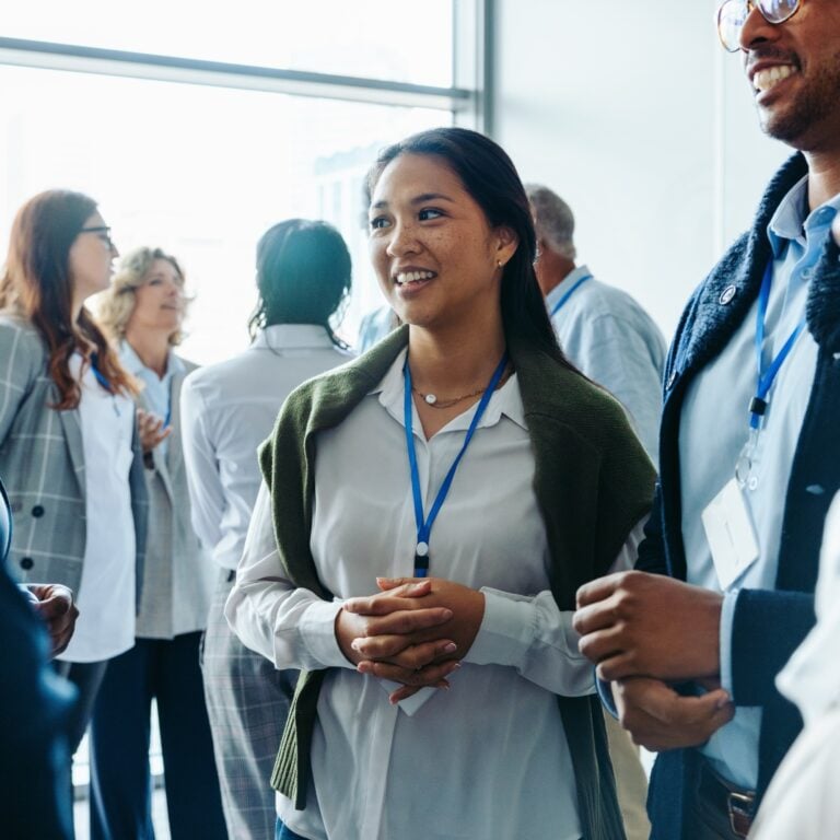 A woman wearing a name badge smiles whilst talking to colleagues at a professional networking event. Several people are chatting in the bright, modern room in the background.