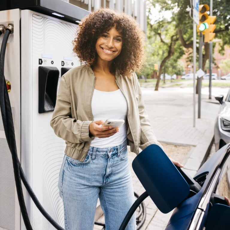 A woman with curly hair smiles while charging her electric car at a charging station outdoors. She holds a mobile phone in one hand and stands next to the car with the charging cable plugged in.