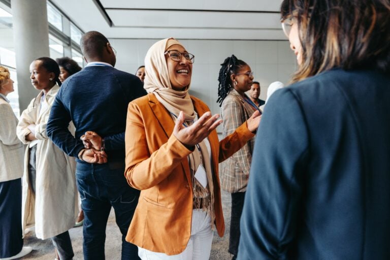 A woman in a tan blazer and hijab smiles and gestures whilst talking to another person at a networking event, with several people engaged in conversation in the background.