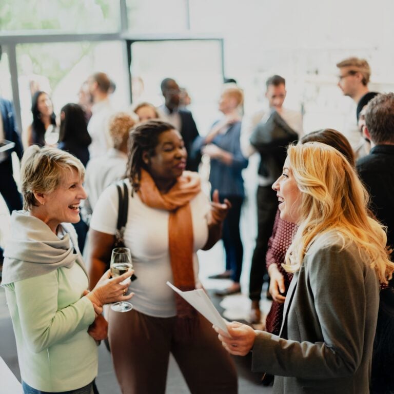 A group of people socialise indoors at a networking event. Two women in the foreground smile and chat, one holding a glass of wine, whilst others converse in the background.