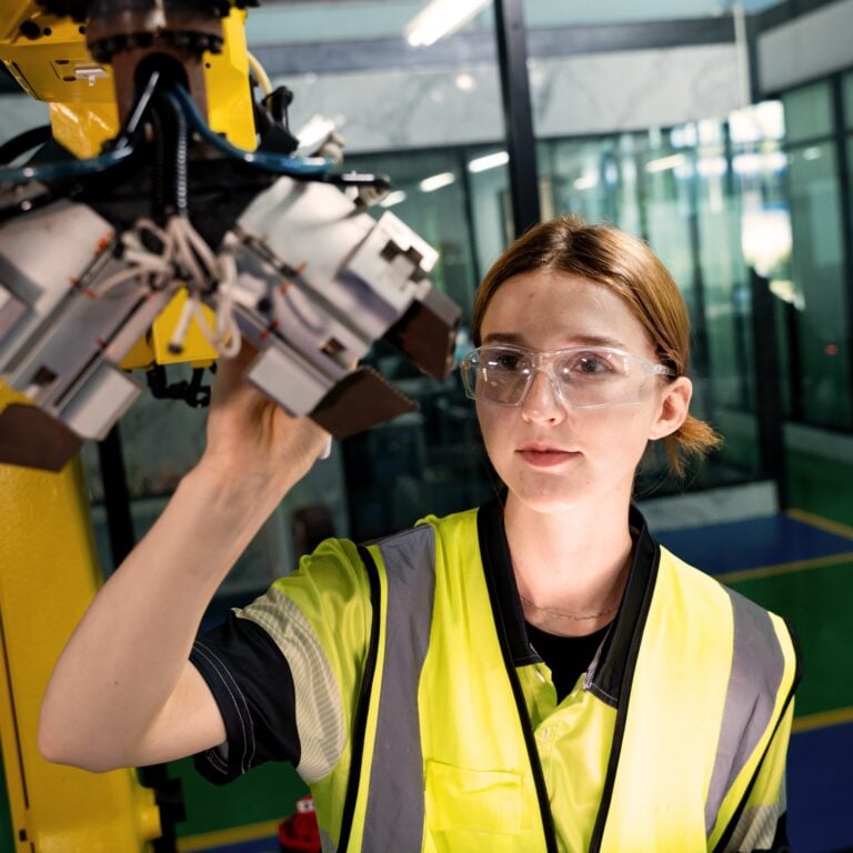 A woman wearing safety glasses and a yellow reflective vest inspects or adjusts a robotic machine arm in a modern industrial setting with glass walls and blue flooring.